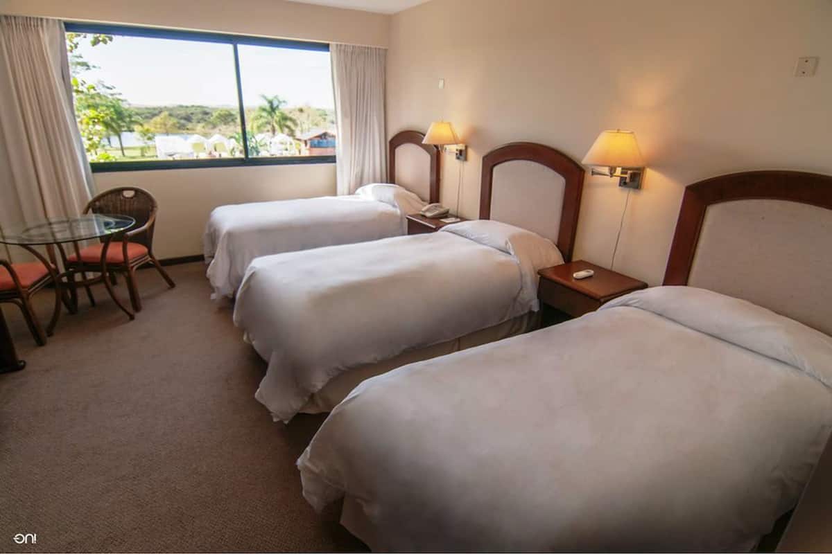 Quarto de hotel com duas camas de solteiro, uma mesa pequena com uma cadeira e uma janela com cortinas.