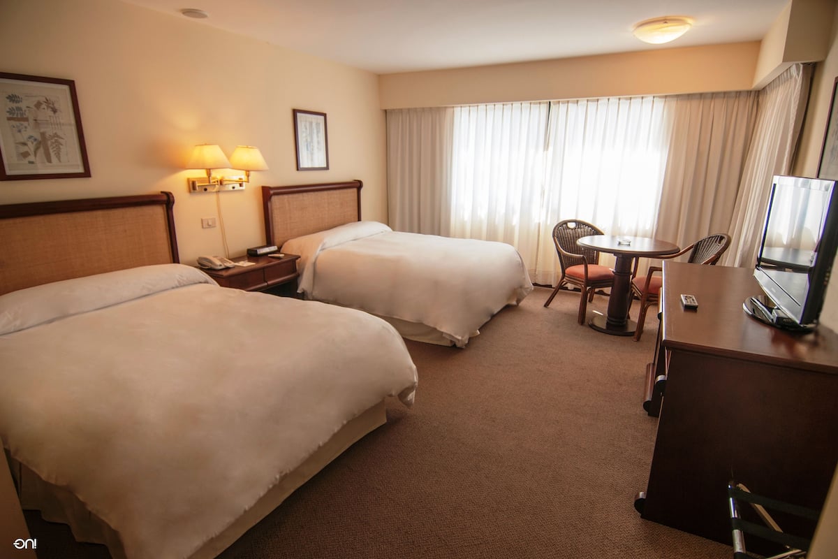 Quarto de hotel com duas camas, uma escrivaninha com televisão, uma cadeira e uma mesa.