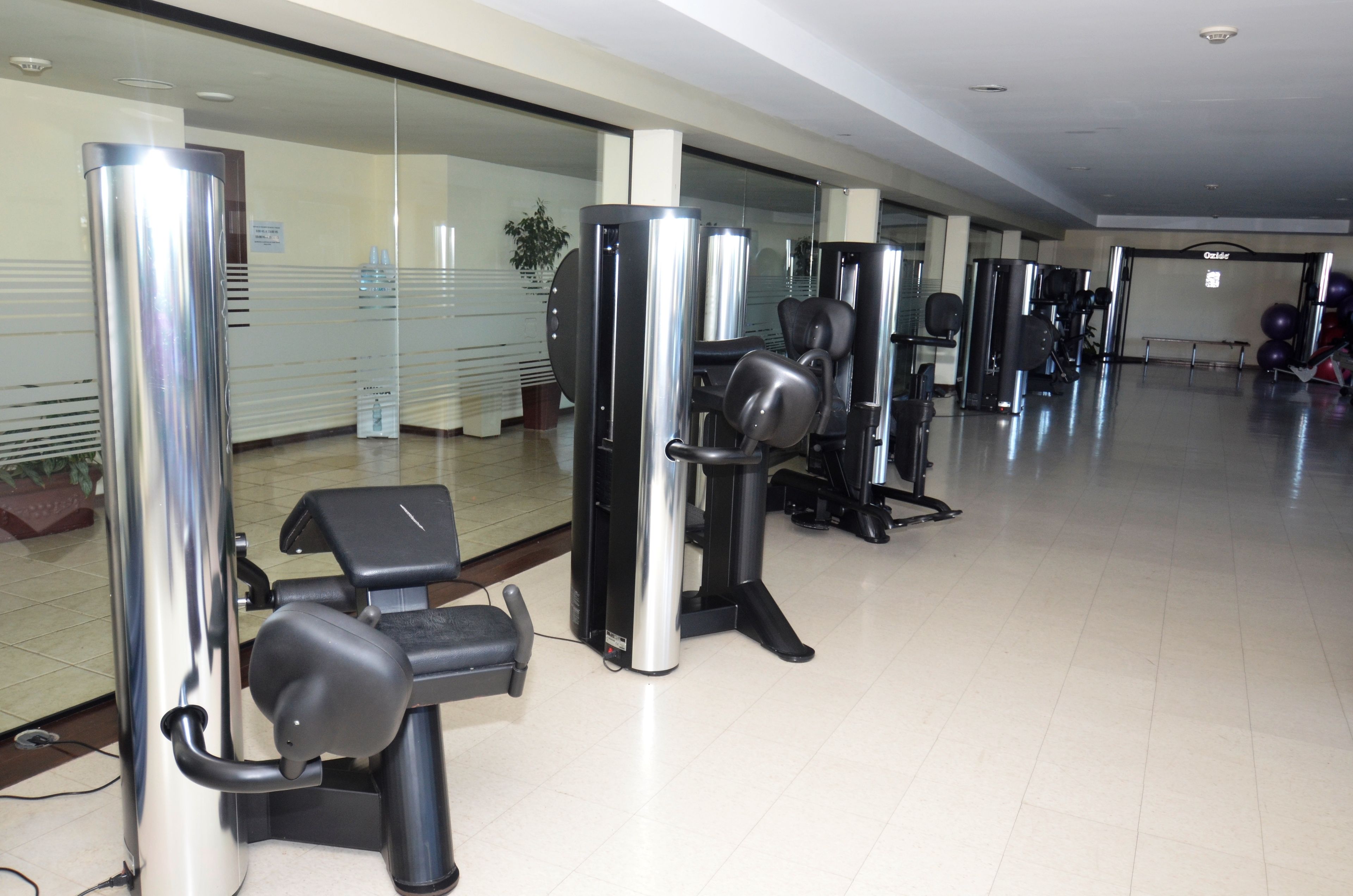 Un gimnasio con diferentes máquinas de ejercicio, un muro de vidrio y piso de baldosas.
