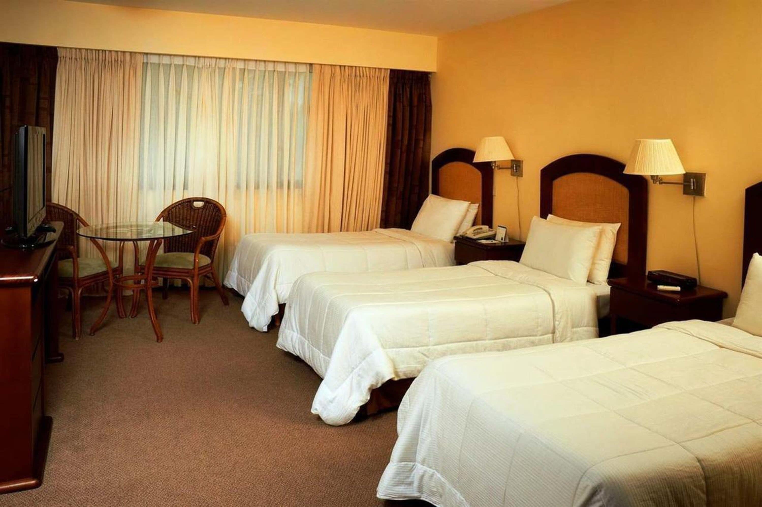 Een hotelkamer met drie enkele bedden, een bureau met stoel, een televisie en een raam met gordijnen.