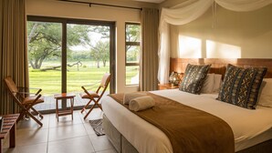 A hotel room with a large bed, two chairs, a small table, and a view of a grassy area through large windows.