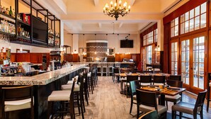 A bar area with a marble counter, multiple bar stools, and a variety of bottles and glasses. There are tables and chairs arranged for dining, and a large window with curtains on the right side. - Royal Park Hotel (Rochester)