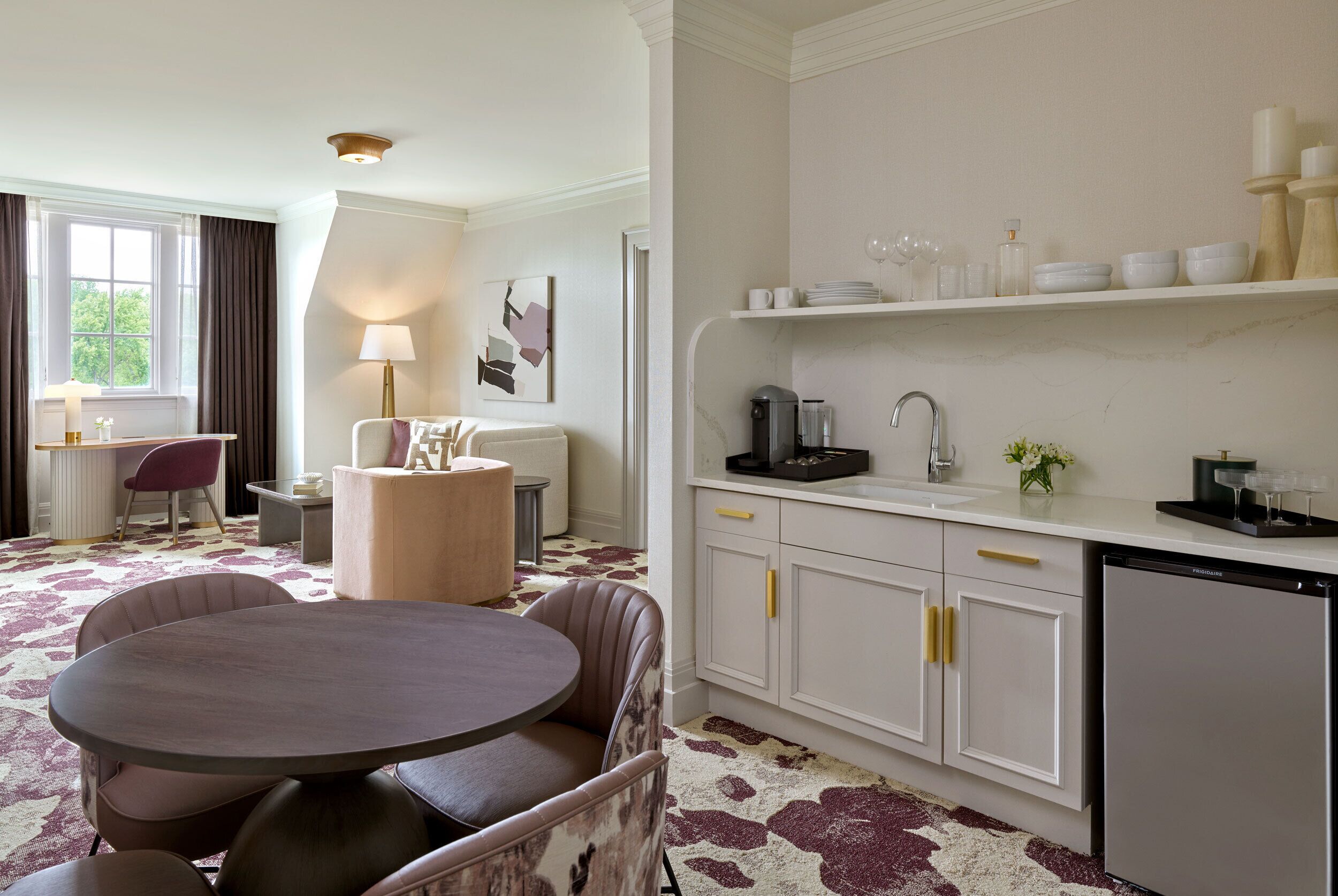 A modern hotel room with a round dining table, a sofa, a kitchen area with a sink and cabinets, and a window with curtains.