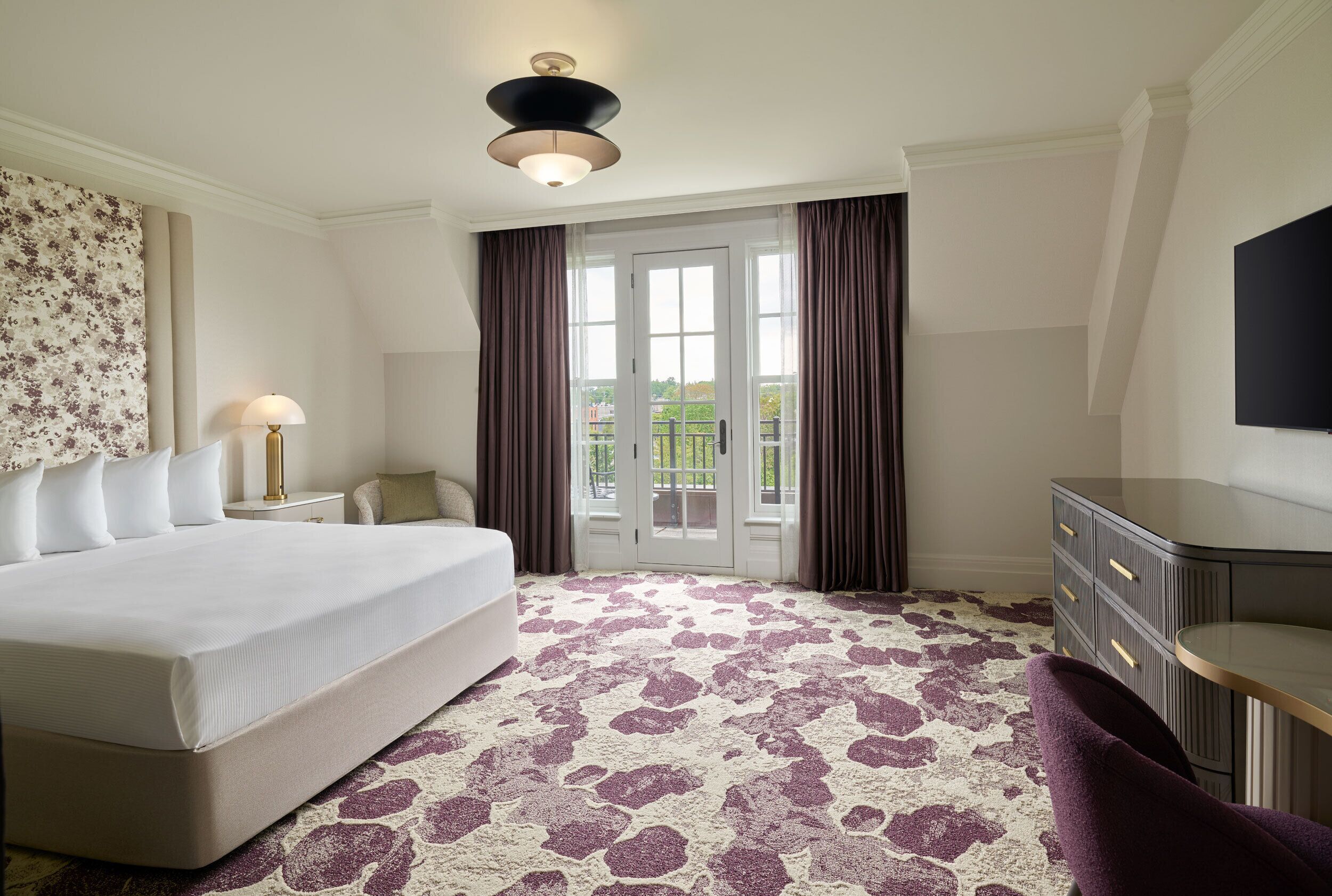A hotel room with a bed, a desk, a chair, a lamp, and a view of the outdoors through a door.