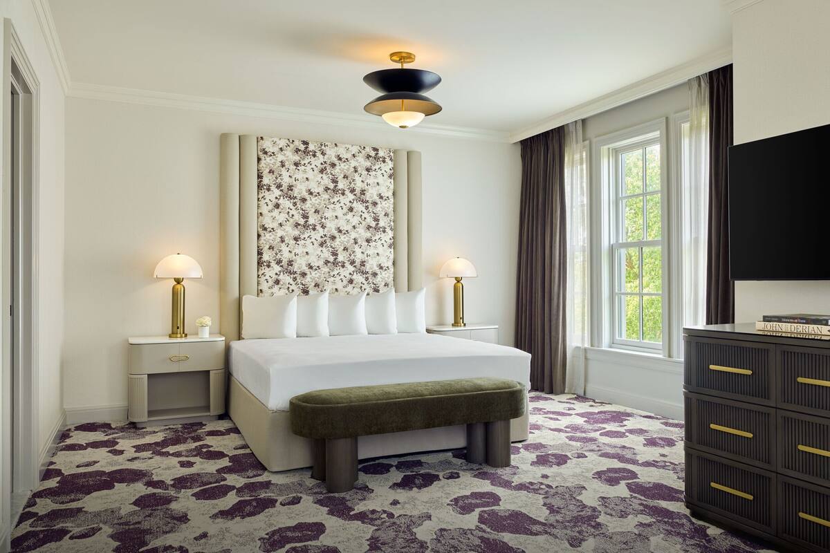 A hotel room with a large bed, two bedside lamps, a bench, a dresser, and a large window with curtains.