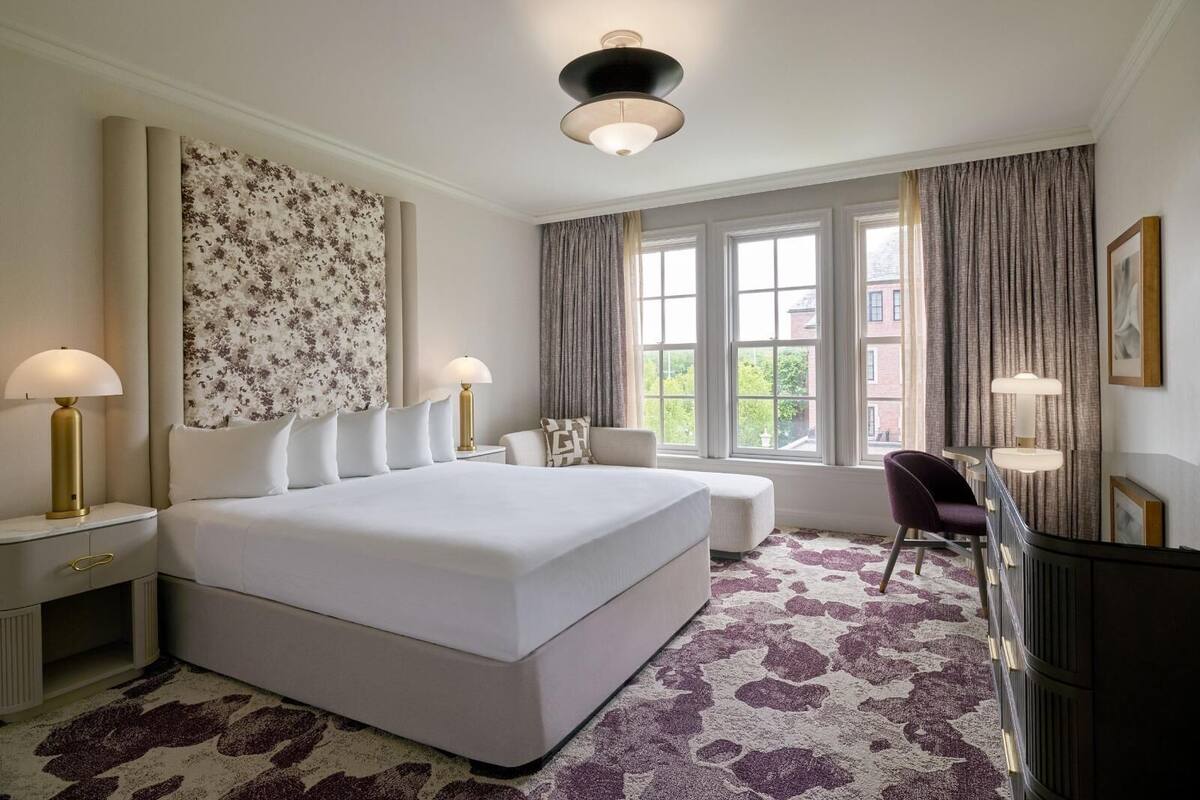 A hotel room with a large bed, two bedside tables with lamps, a desk with a chair, and a large window with curtains.