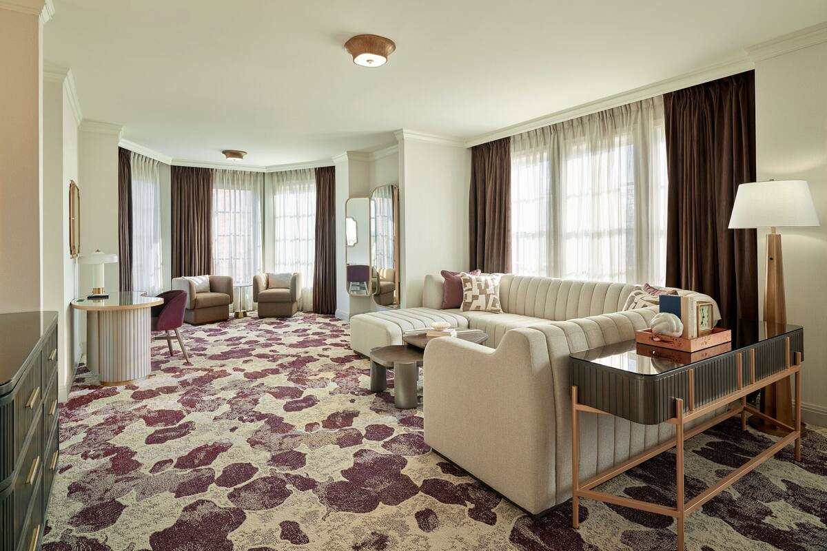 A spacious living room with a large sofa, a coffee table, and a TV. There is a dining area with a table and chairs, and a kitchen area with a counter and cabinets.
