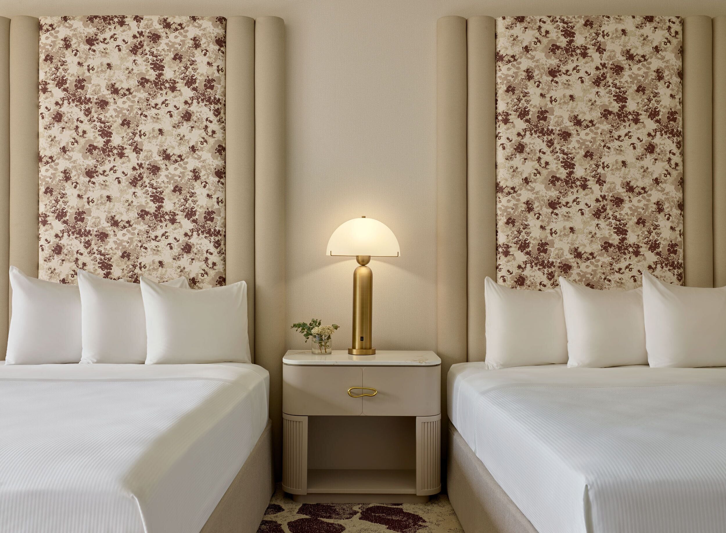 Una habitación de hotel con dos camas, una mesita de noche con lámpara y papel tapiz con motivos florales.