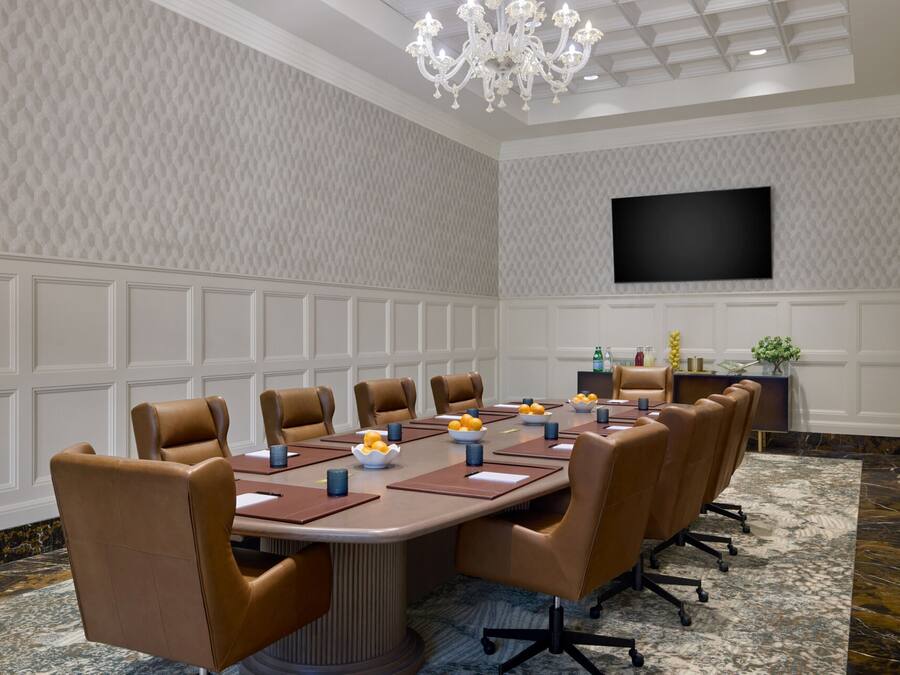 A conference room with a long table, brown leather chairs, a chandelier, a flat-screen TV, and a patterned wall.