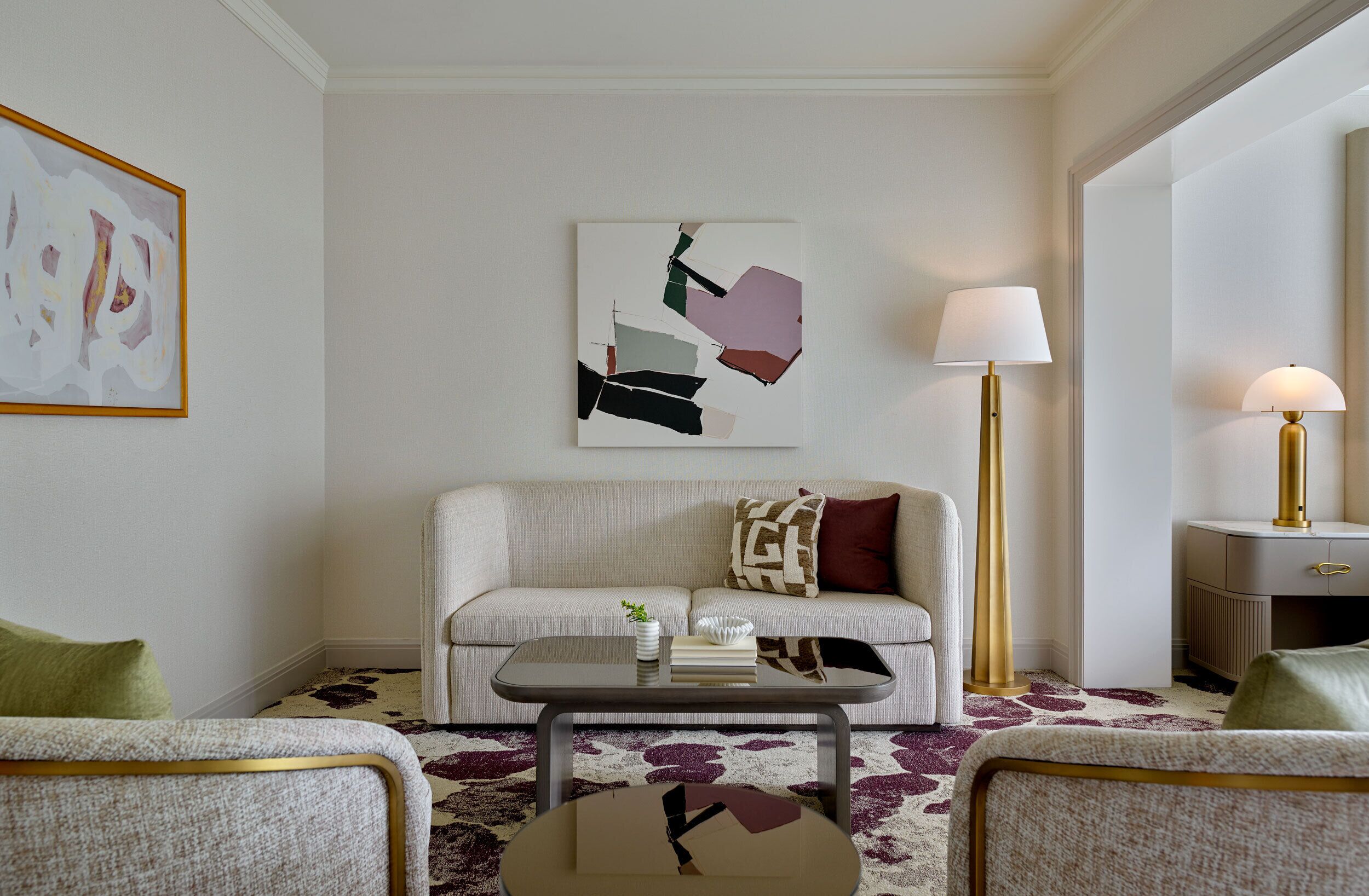 A modern living room with a sofa, two armchairs, a coffee table, and a floor lamp. There is a painting on the wall and a small table with books and a plant.