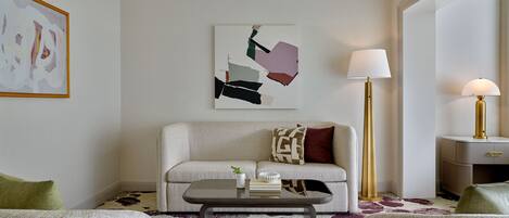 A modern living room with a sofa, two armchairs, a coffee table, and a floor lamp. There is a painting on the wall and a small table with books and a plant.