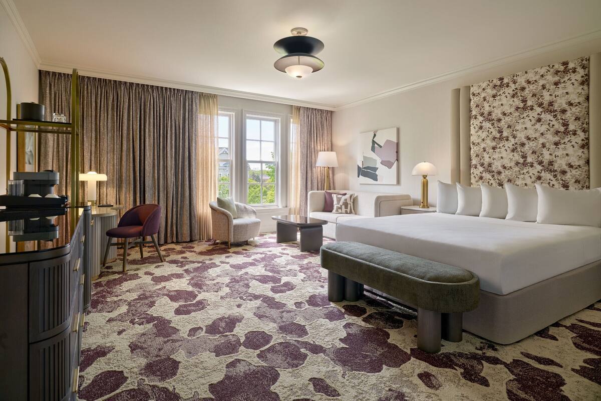 A hotel room with a large bed, a desk with a chair, a sofa, a bench, and a large window with curtains.