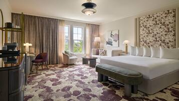A hotel room with a large bed, a desk with a chair, a sofa, a bench, and a large window with curtains.