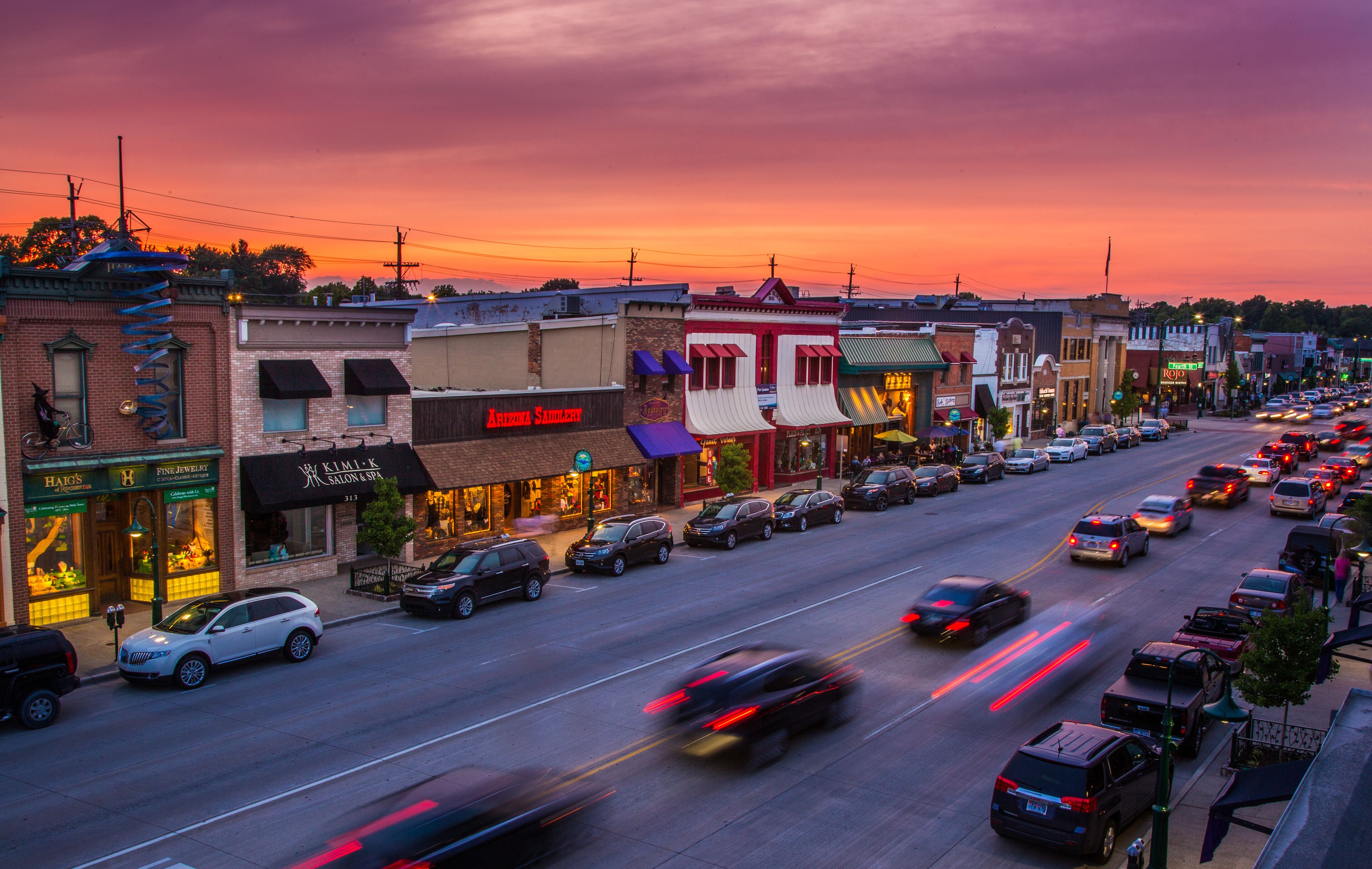 A street lined with various shops and restaurants, with cars parked and driving on the road.