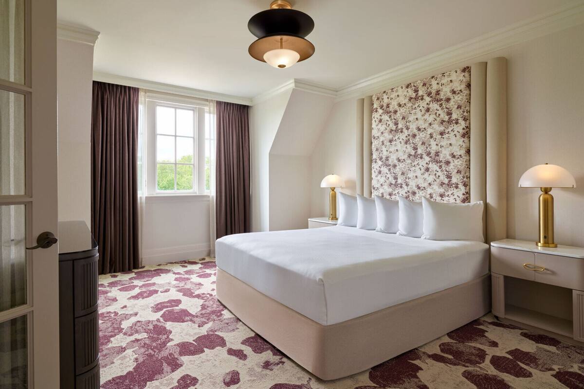 A large bed with a headboard, two bedside tables with lamps, a window with curtains, and a patterned carpet.