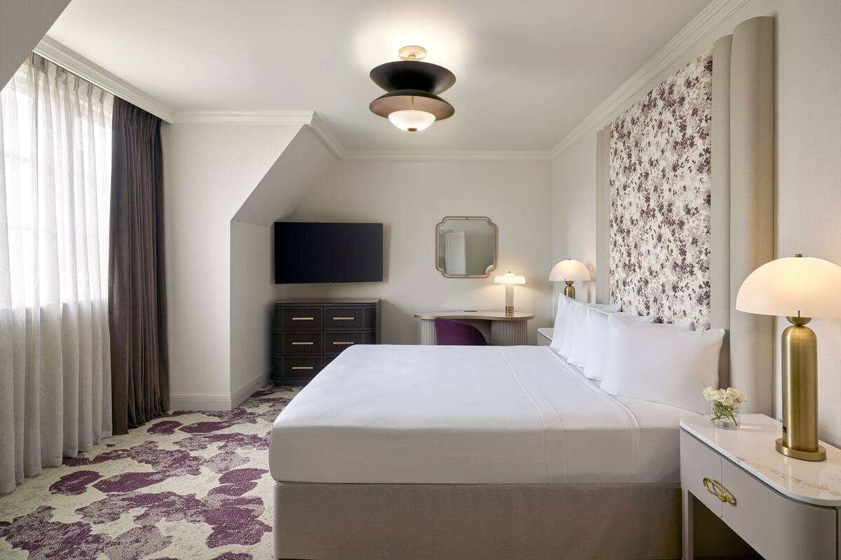 A hotel room with a large bed, a desk with a lamp, a flat-screen TV, and a decorative wall panel.