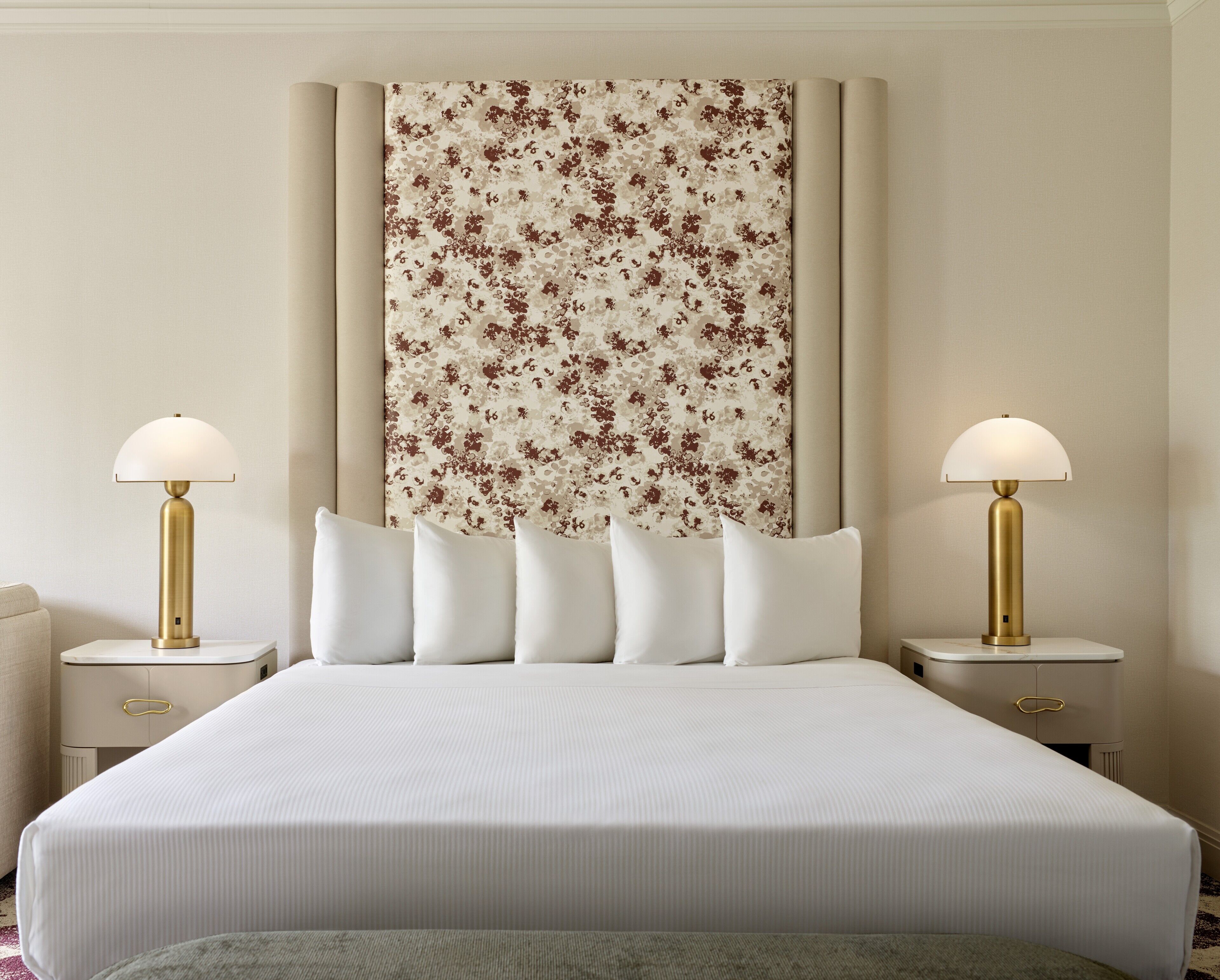 A neatly made bed with a floral headboard, two bedside lamps, and a bedside table with a drawer.