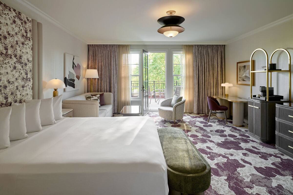 A hotel room with a large bed, a desk, a chair, and a balcony with curtains.