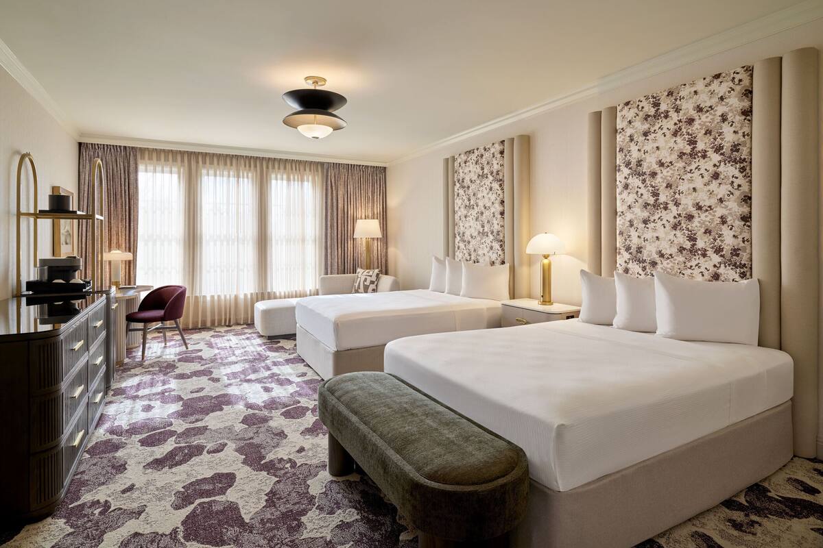 A hotel room with two beds, a desk, a chair, and a large window with curtains.