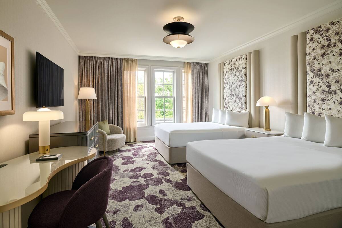A hotel room with two beds, a desk, a chair, and a large window with curtains.