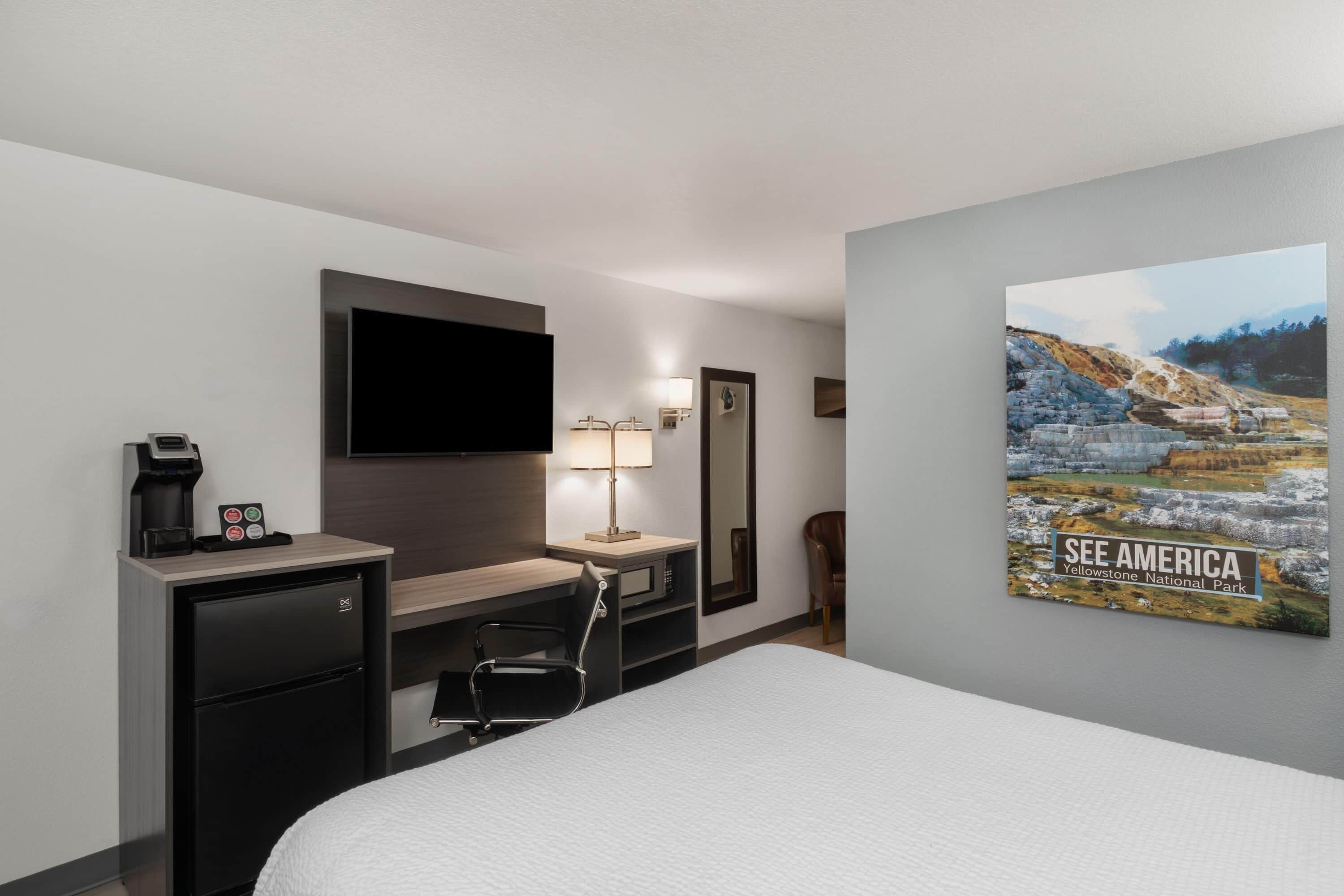 A hotel room with a bed, a desk, a chair, a TV, a coffee machine, and a poster on the wall.