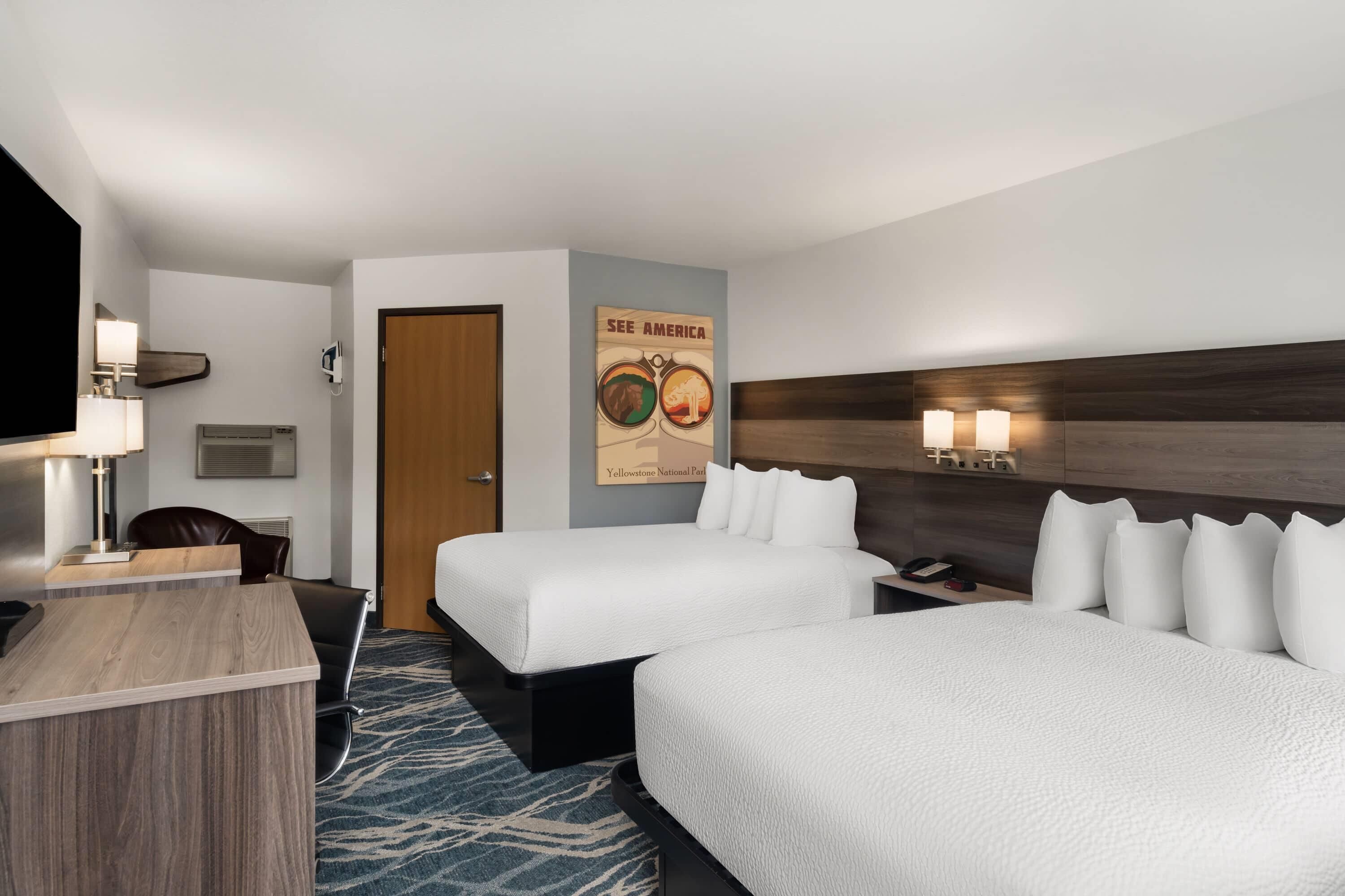 A hotel room with two beds, a desk, a chair, a wall-mounted TV, and a poster advertising Yellowstone National Park.