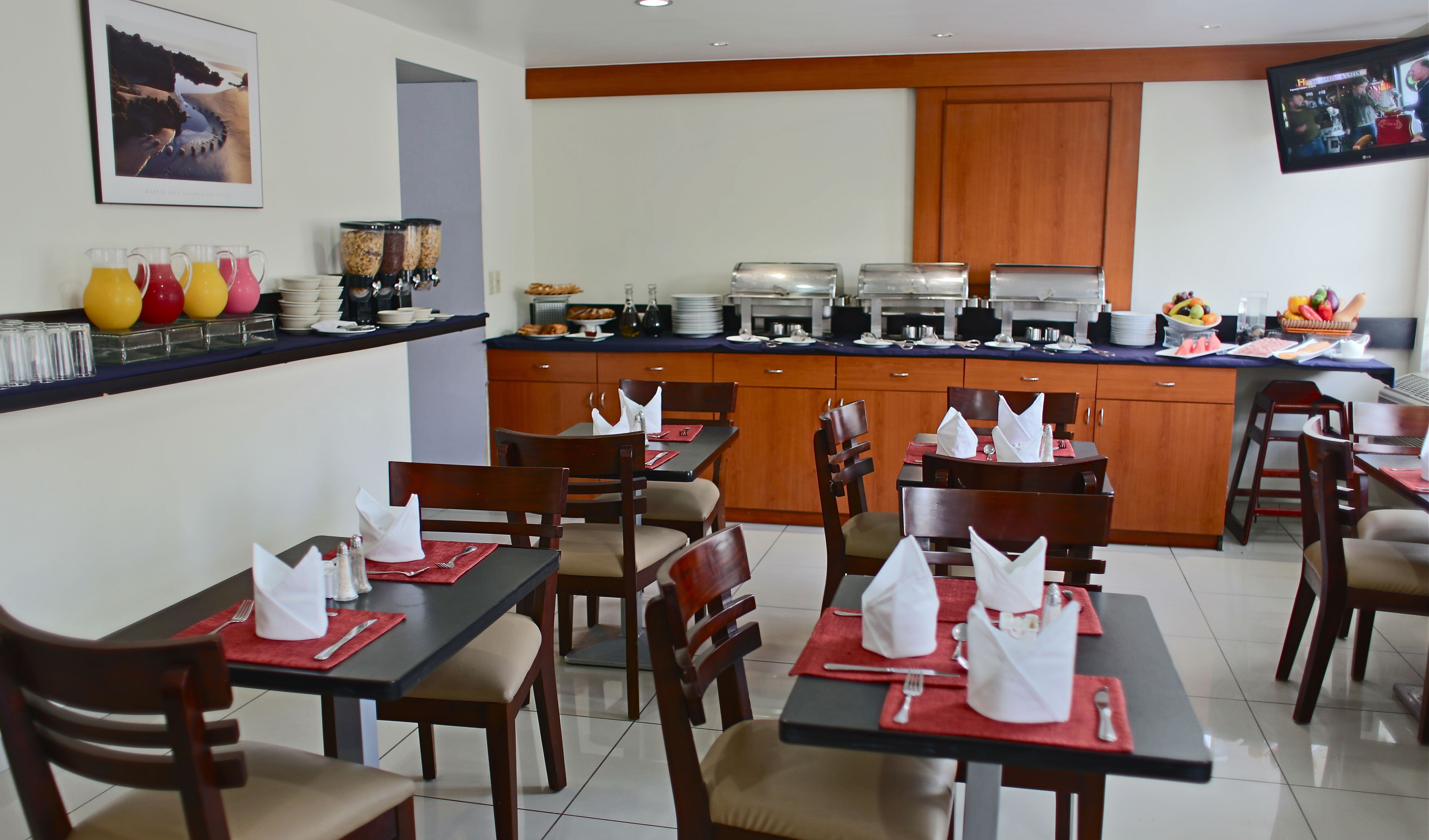 A dining area with tables set for guests, a buffet counter with various food items, and a television mounted on the wall.
