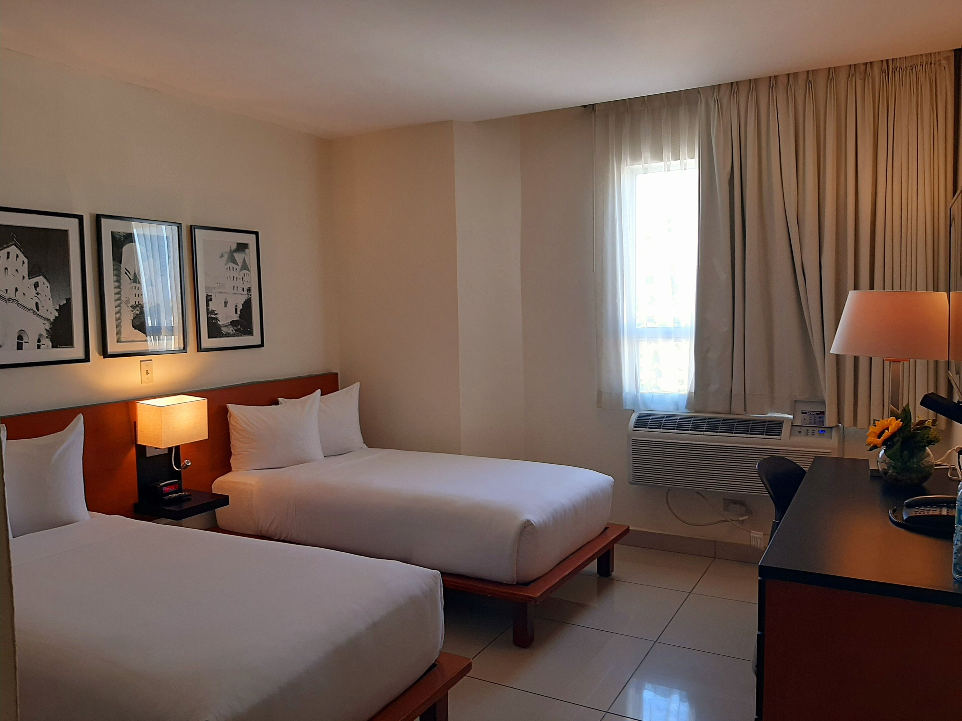 A hotel room with two beds, a desk, a lamp, a vase with flowers, and a window with curtains.