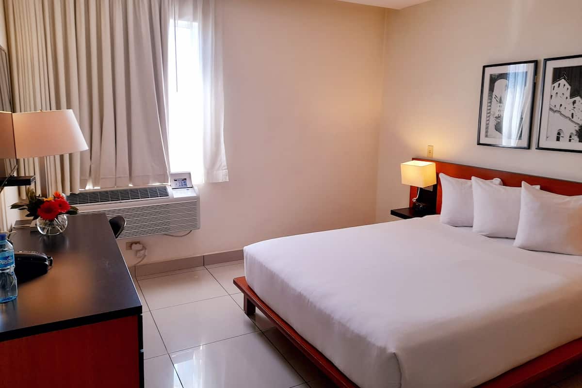 A hotel room with a bed, bedside lamps, a desk with a lamp and a vase of flowers, a window with curtains, and two framed pictures on the wall.