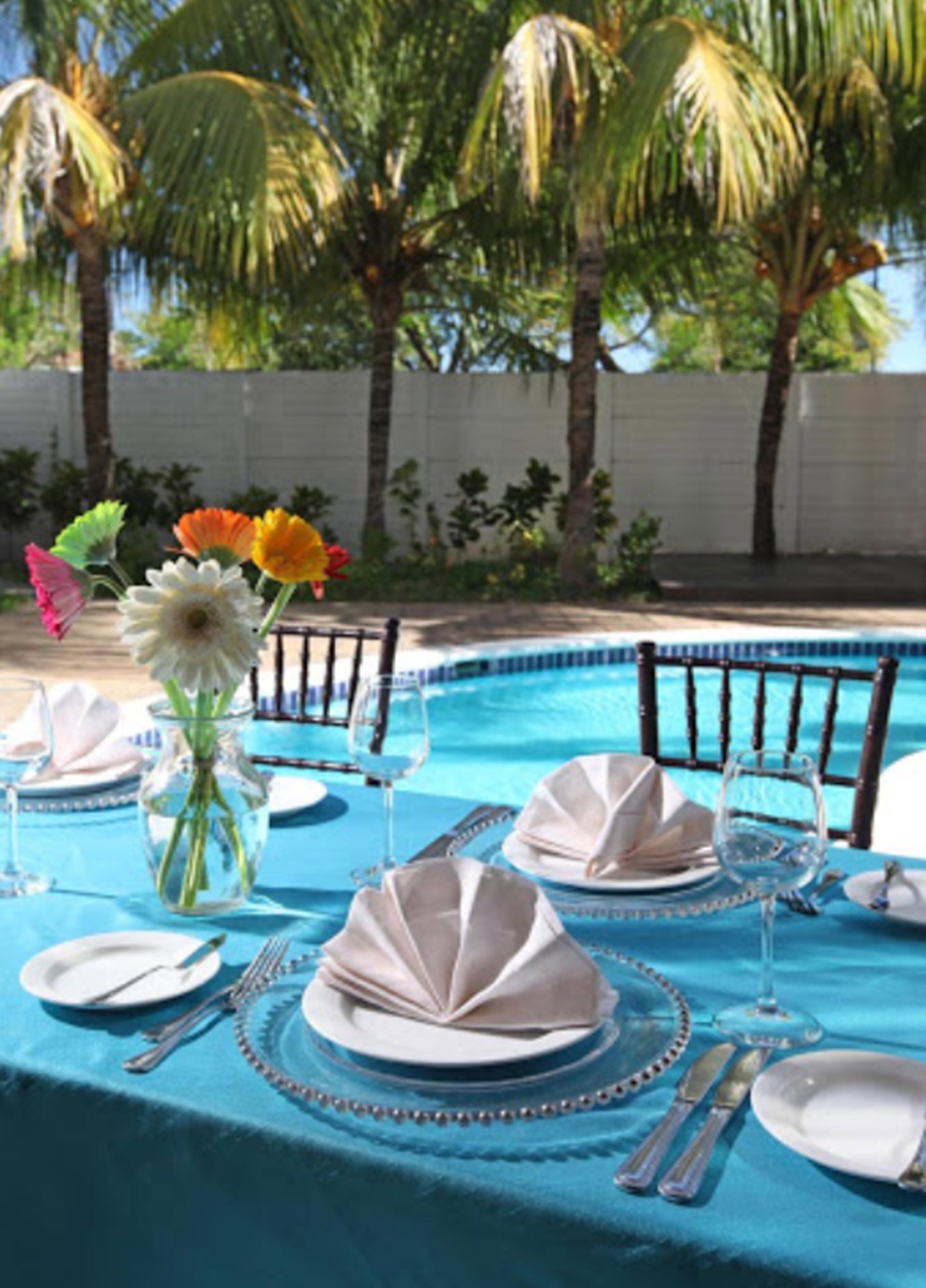 A poolside dining area with a blue tablecloth, white table settings, and a vase of flowers.