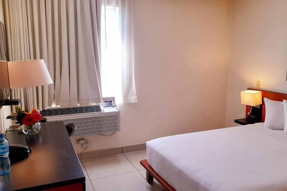 A hotel room with a bed, a desk with a lamp, a water bottle, a radiator, and curtains.