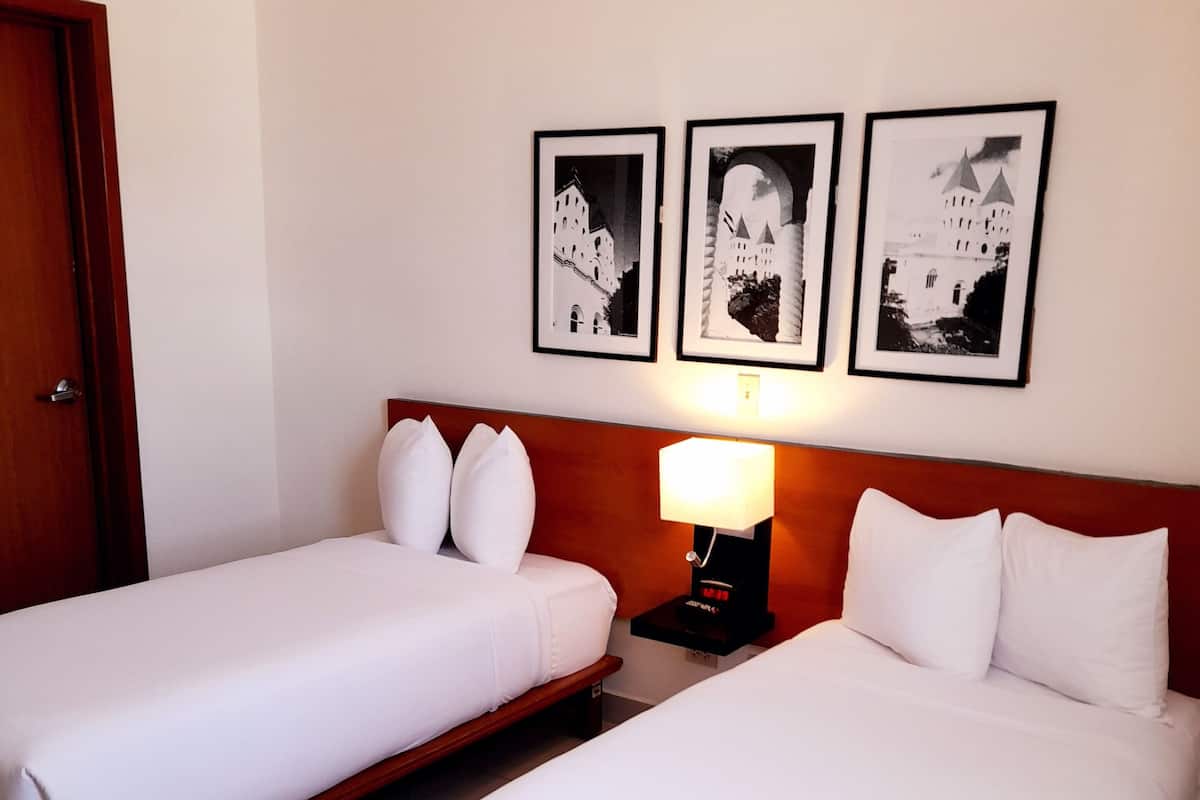 A hotel room with two single beds, a wooden headboard, a nightstand with a lamp, and three framed pictures on the wall.