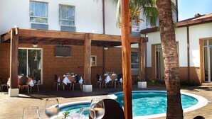 Un área de comedor junto a la piscina con mesas y sillas, piscina y un porche cubierto.