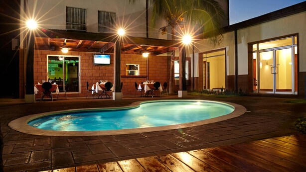 Zona de piscina con patio de ladrillo, mobiliario exterior y un edificio con pantalla de televisión y zona de bar.