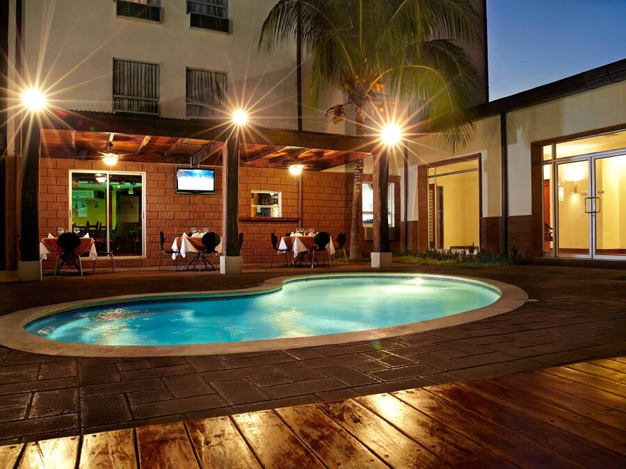 A pool area with a brick patio, outdoor seating, and a building with a TV screen and a bar area.