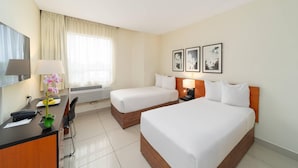Habitación de hotel con dos camas, un escritorio con lámpara, una silla, televisión y cuadros en la pared.