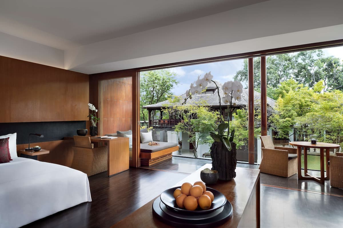 A modern hotel room with a large bed, a dining area, and a view of a traditional building and trees.