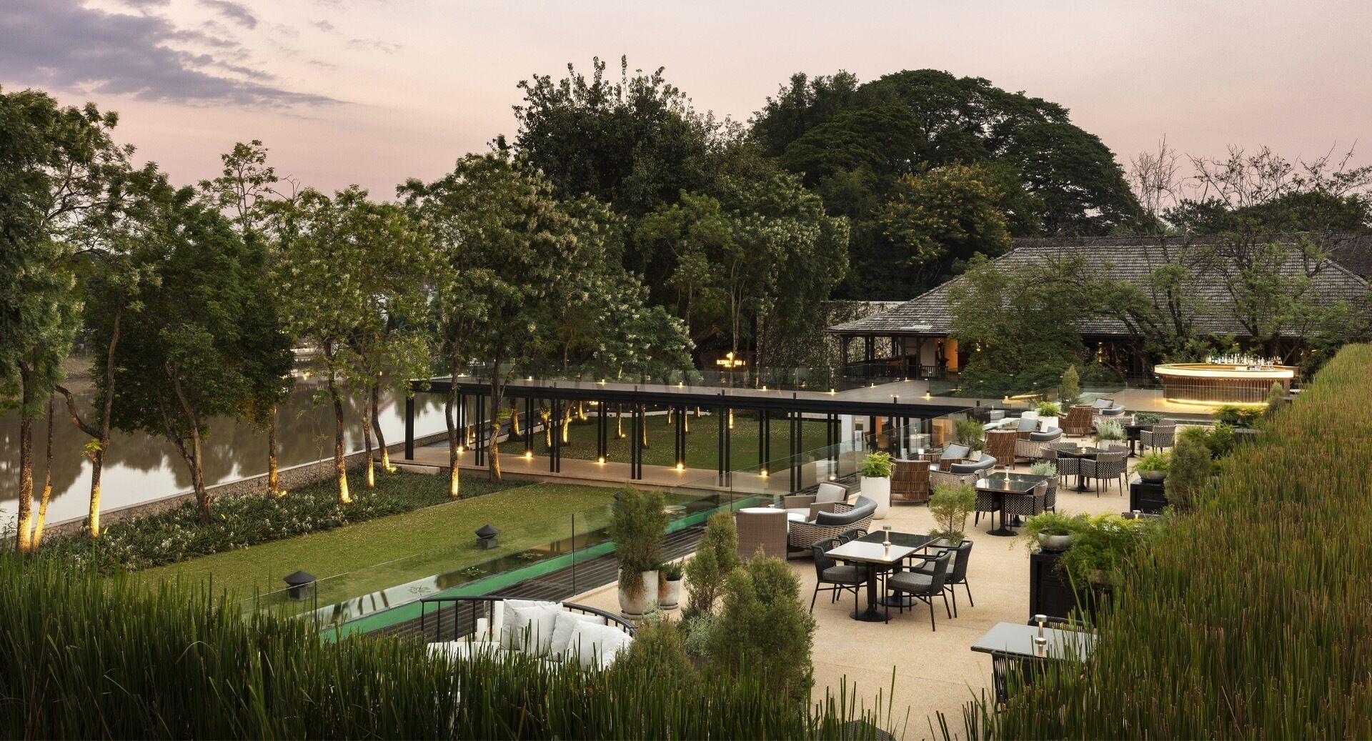 A modern outdoor dining area with tables and chairs, surrounded by lush greenery and a body of water.