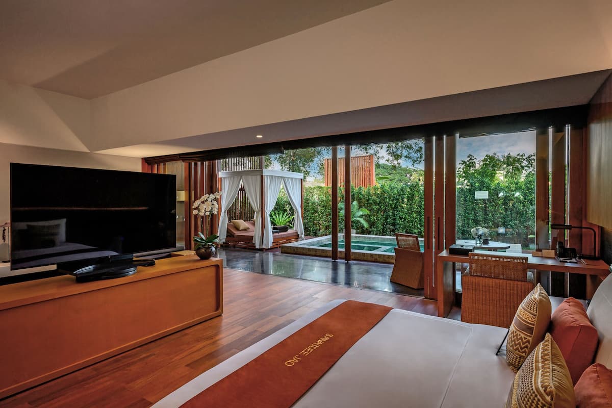 A modern hotel room with a large TV, a comfortable sofa, a dining area, and a view of the garden.