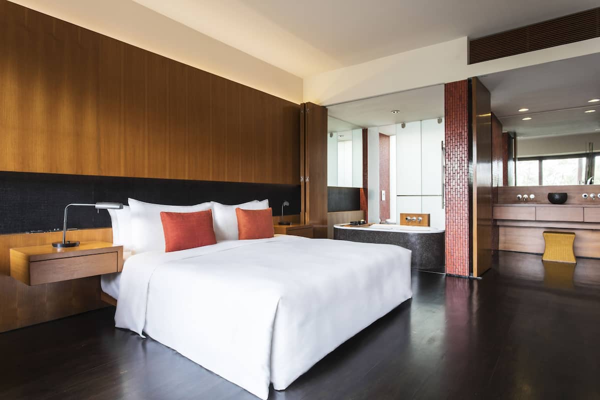 A modern hotel room with a large bed, a wooden headboard, a nightstand with a lamp, a bathroom with a bathtub, and a large mirror.