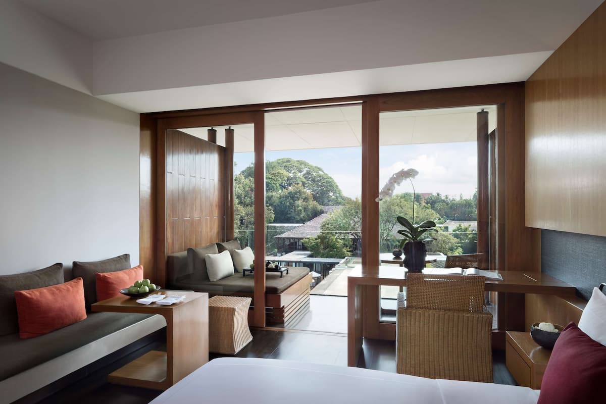 A modern hotel room with a sofa, a dining table, and a chair. There is a large window offering a view of the outdoors.