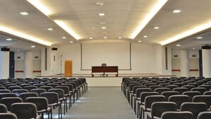 Un salón de conferencias con filas de sillas, un atril, una pantalla y luces en el techo.