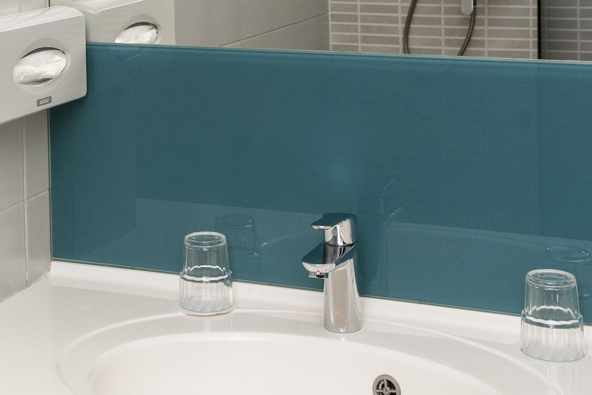 Un baño con un lavamanos blanco, dos vasos de vidrio, un cabezal de ducha de mano y un perchero para toallas.