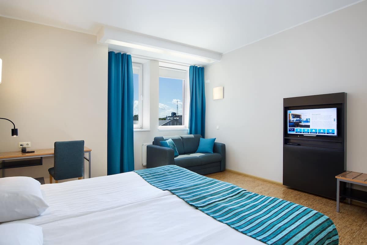 A hotel room with a bed, a desk, a chair, a TV, and a window with curtains.