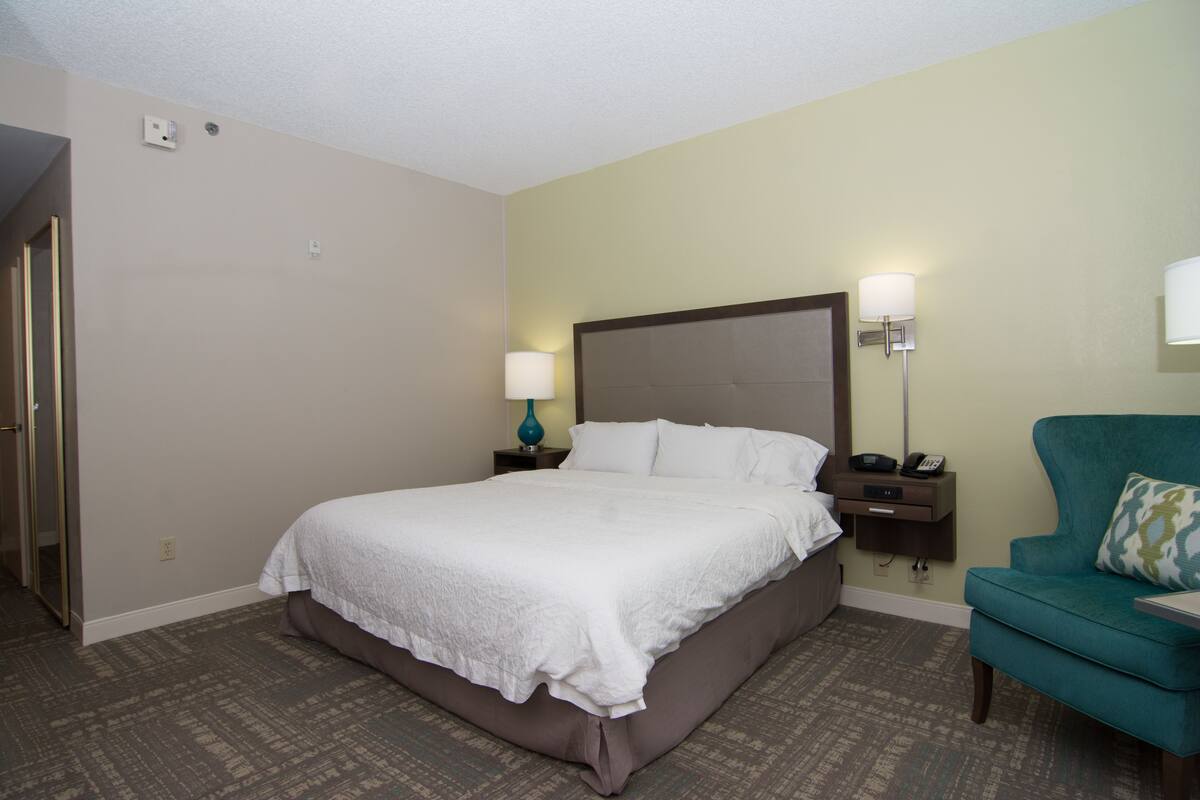 A hotel room with a large bed, two bedside tables with lamps, a teal armchair, and a patterned carpet.