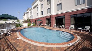 Una piscina con sillones de descanso y sombrillas, rodeada por un patio de ladrillo y un edificio con ventanas grandes.