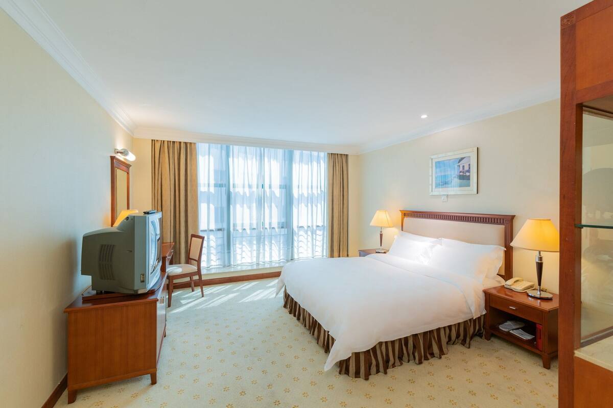 A hotel room with a large bed, a television on a wooden stand, a desk with a lamp, and a window with curtains.