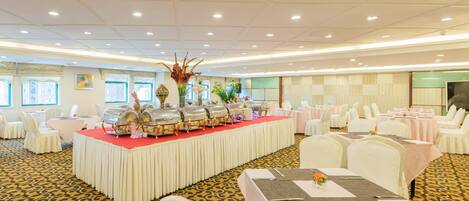 Une salle de banquet avec des tables rondes, des chaises blanches et un espace buffet où sont disposés des plats.