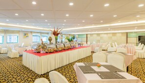Un salón de banquetes con mesas redondas, sillas blancas y una zona de bufé con recipientes de comida.