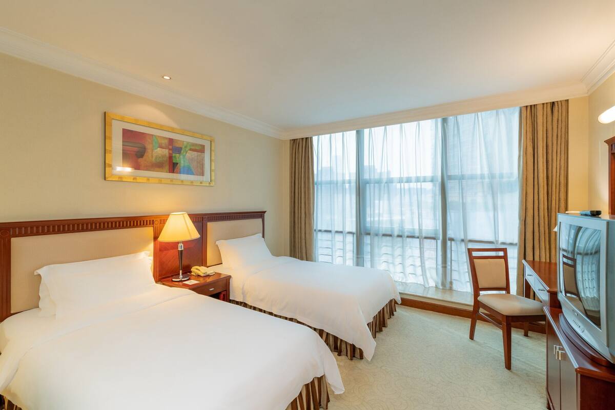 A hotel room with two beds, a desk with a lamp, a television, and a large window with curtains.